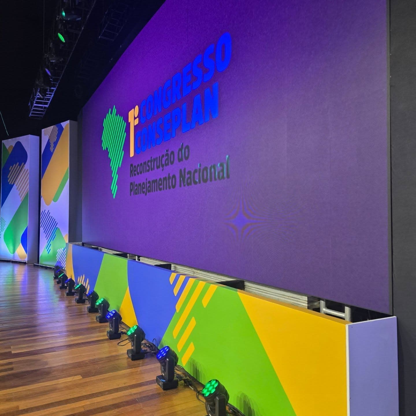 Vista lateral do palco de congresso com painel de LED e linha de monitores.