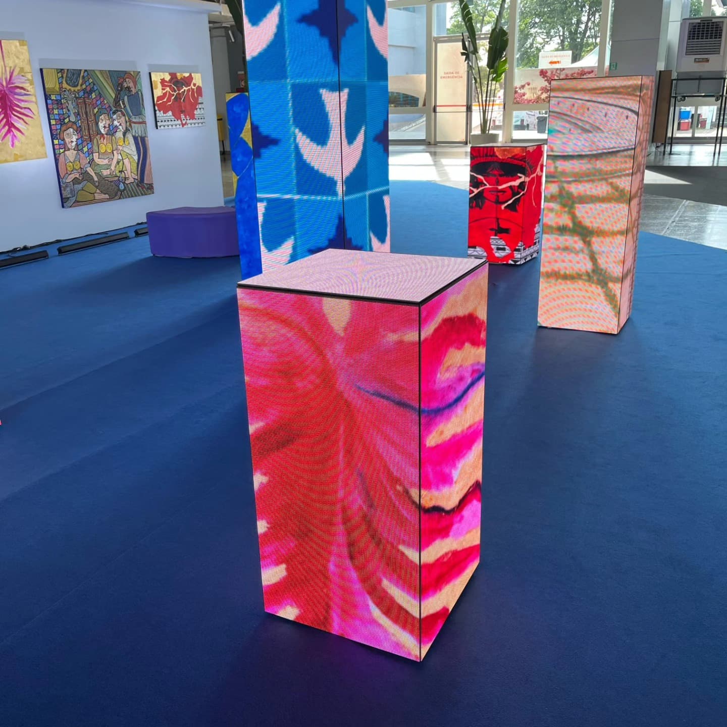 Cubos e totens de painel de LED exibindo obras coloridas em um espaço expositivo.